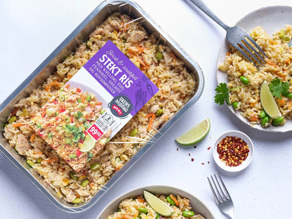 Stekt ris i en aluminiumsform med grønnsaker og krydder, ser ut til å være en ferdiglaget middag, med limebåter og krydder ved siden av.