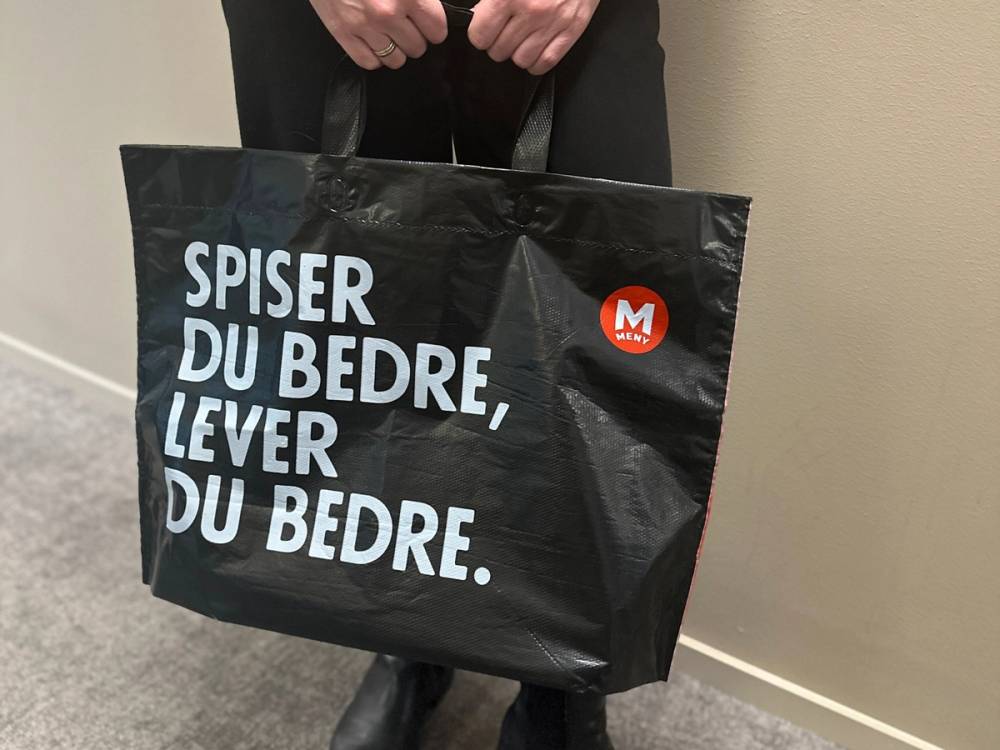 Person holder en svart pose med teksten 'Spiser du bedre, lever du bedre' og en liten rød sirkel med hvit M inni, mot en beige vegg.