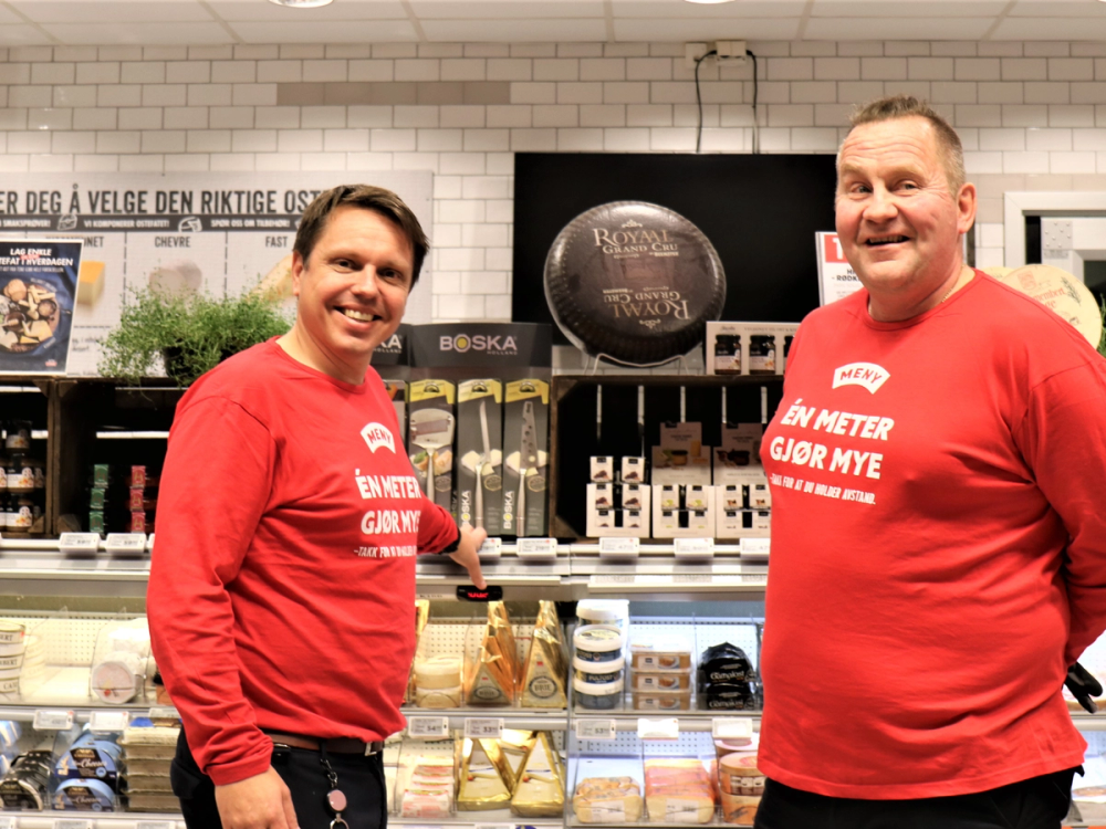 To personer i røde klær står i en butikk med hyller fylt med ulike produkter, og en TV-skjerm henger på veggen bak dem.