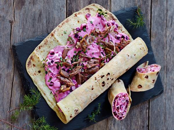 Pinnekjøtt-lefse med rosa kål og sopp på en svart tallerken, dekorert med dill, på en treplate.