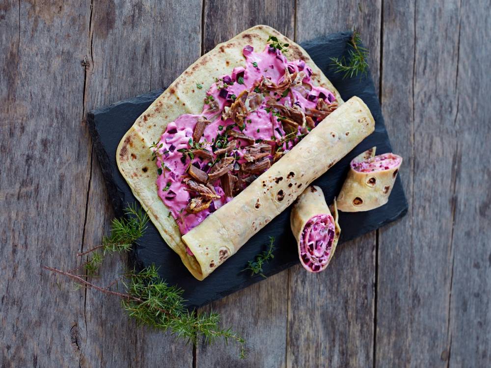 Pinnekjøtt-lefse med rosa kål og sopp på en svart tallerken, dekorert med dill, på en treplate.