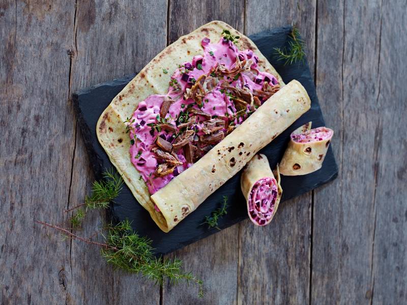 Pinnekjøtt-lefse med rosa kål og sopp på en svart tallerken, dekorert med dill, på en treplate.