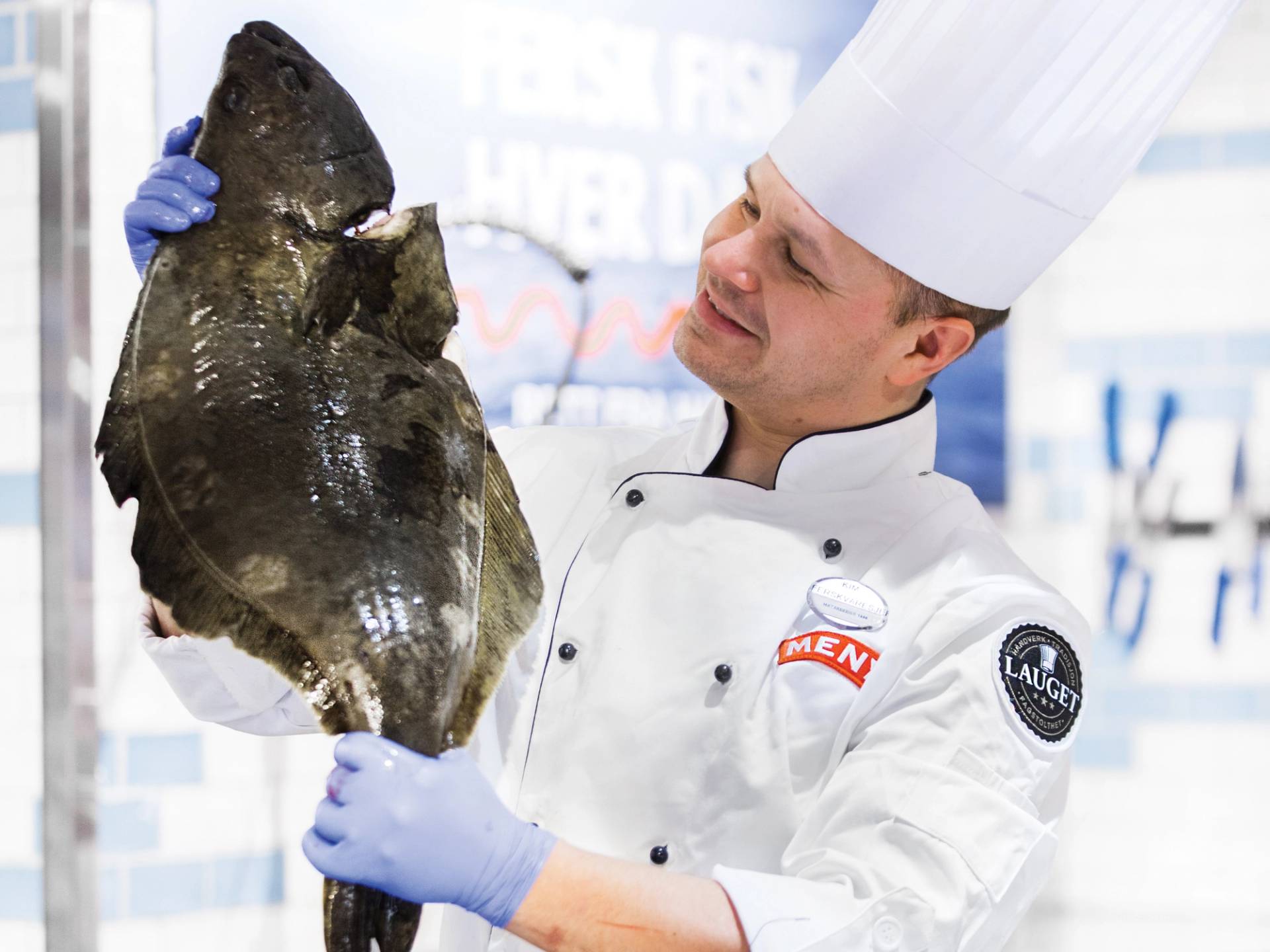 En kok i hvit kokkeuniform og lue holder opp en stor, mørk kveite under en utstilling eller konkurranse.