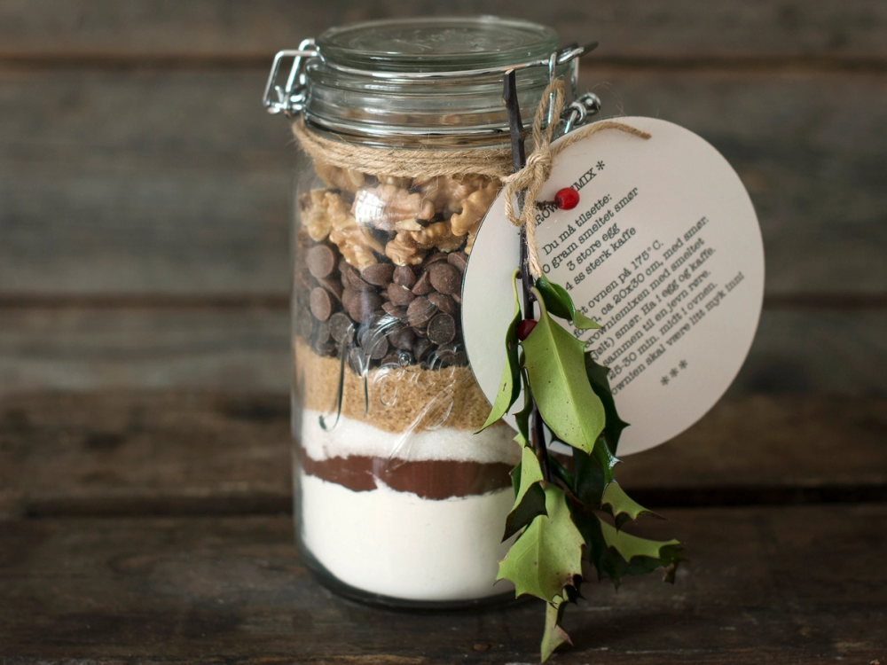 Et glass med lag av brownie, sjokoladebiter, og krem, dekorert med en kvist med grønne blader og en etikett, på en treoverflate.