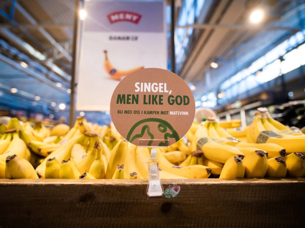 Bananer i en fruktdisplay med en plakat som sier 'Singel, men like god' på MENY butikken, med butikkhyller og belysning i bakgrunnen.