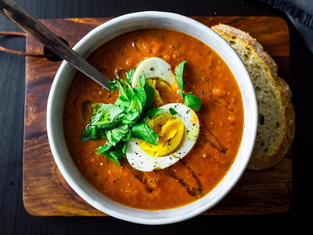 Tomatsuppe med skiver av hardkokt egg og fersk basilikum i en hvit bolle, med brødskiver ved siden av.