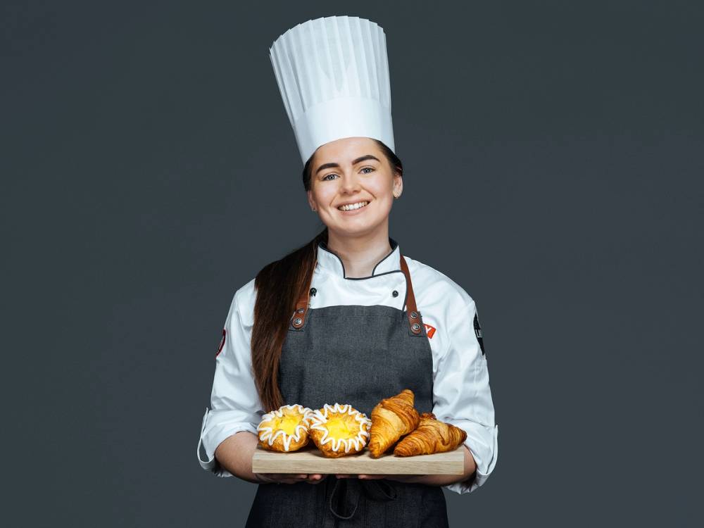 En person kledd som baker, iført kokkehatt og forkle, holder et brett med kaker og croissanter.