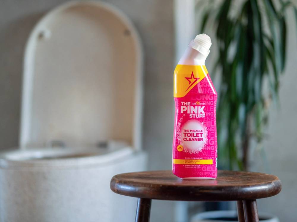 En flaske med rengjøringsmiddel kalt The Pink Stuff står på en liten trebord foran en åpen toalettlokk, med grønne planter i bakgrunnen.