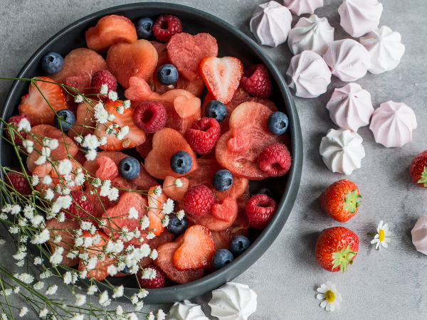 En skål med ulike frosne bær og vannmelonbiter, omgitt av små hvite blomster og noen jordbær med grønne stilker.