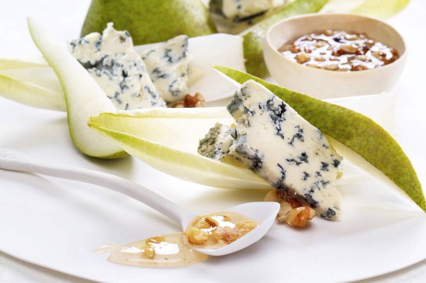 Blåmuggost med pære og honning på et hvitt fat, omgitt av flere pærer og en skål med syltetøy eller saus.