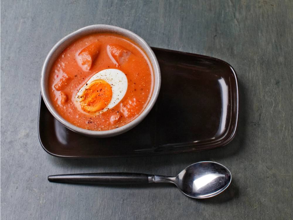 En kopp med tomatsuppe med en skive hardkokt egg oppå, plassert på en svart tallerken med en skje ved siden av, på et grått bord.