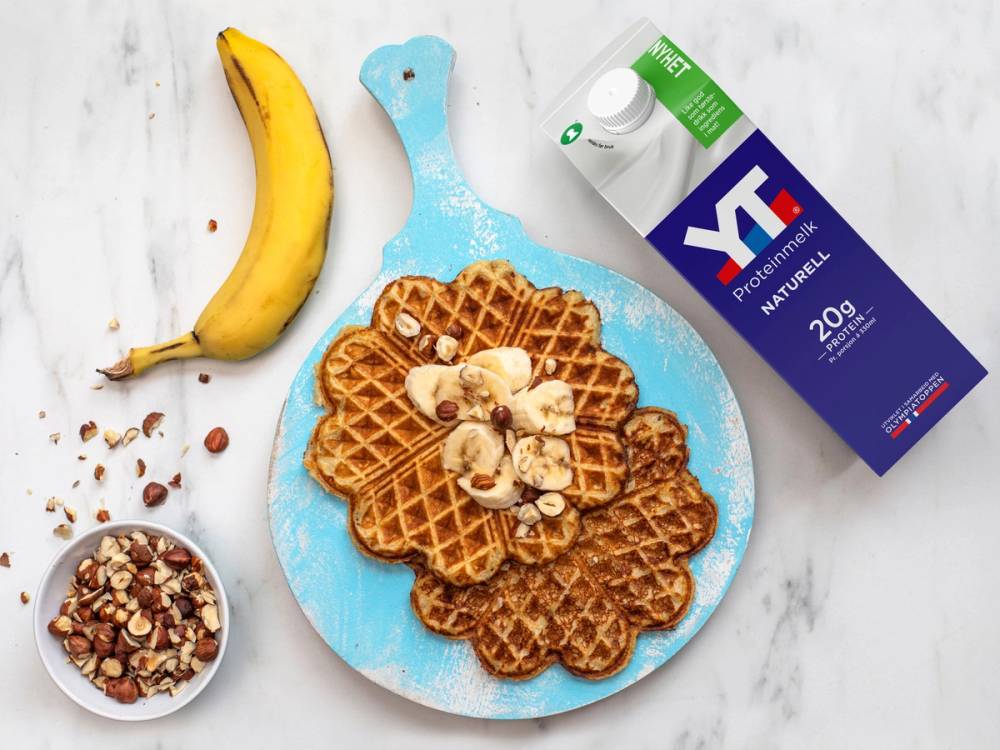 Proteinvaffel med banan og havregryn på en blå serveringsplate, sammen med en banan, en kartong TINE PROTEINMELK og en liten skål med nøtter og frø.