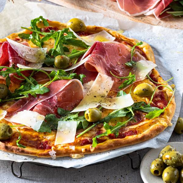 Pizza med parmaskinke, grønne oliven, ruccola og høvlet parmesanost på toppen.
