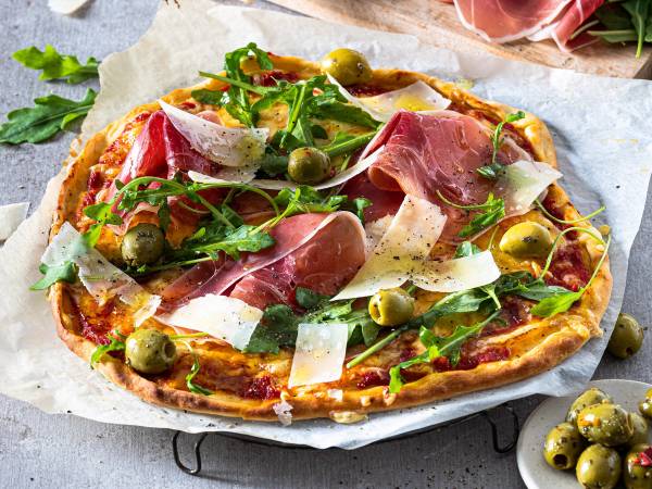 Pizza med parmaskinke, grønne oliven, ruccola og høvlet parmesanost på toppen.