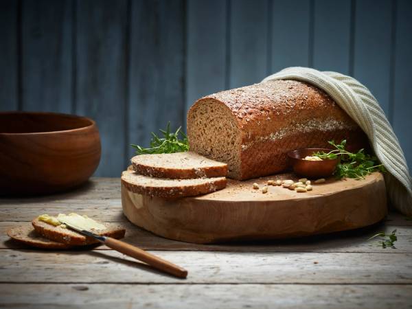 Et stort, grovt brød ligger på et skjærebrett med noen skiver skåret av, omgitt av en liten skål med smør og noen urter, på et mørkt trebord med en blå bakgrunn.