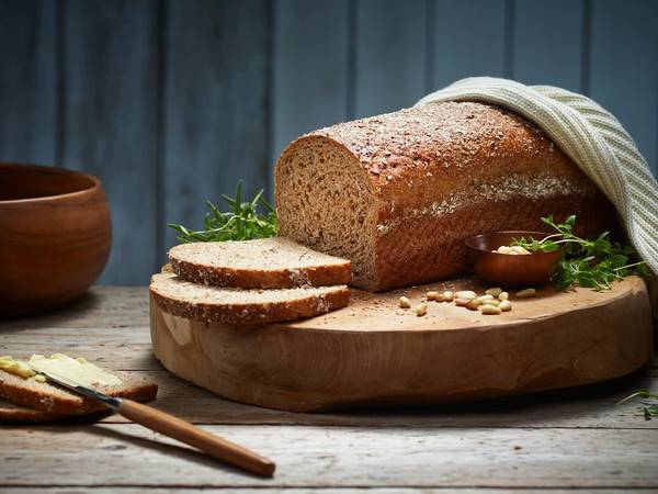 Et stort, grovt brød ligger på et skjærebrett med noen skiver skåret av, omgitt av en liten skål med smør og noen urter, på et mørkt trebord med en blå bakgrunn.