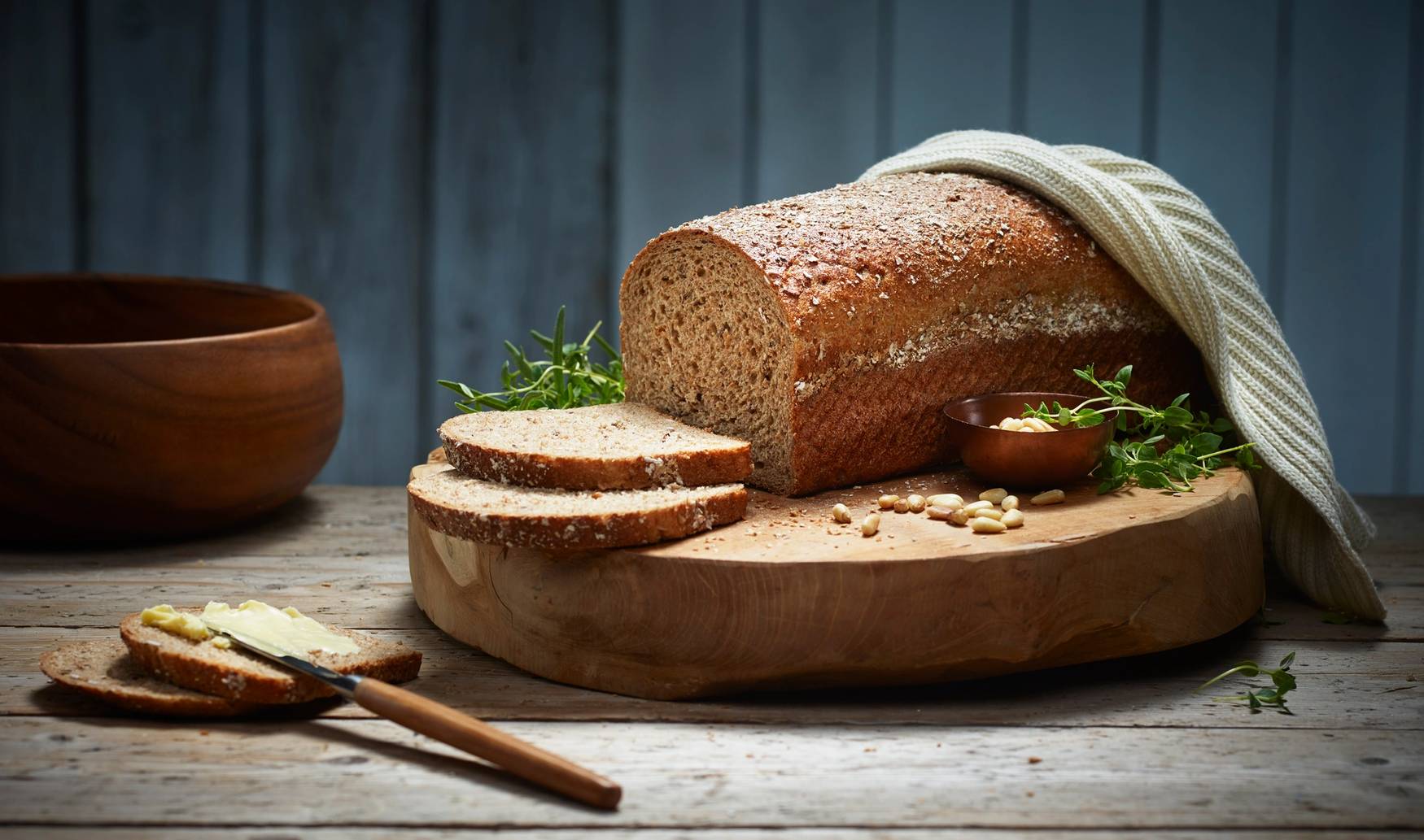Et stort, grovt brød ligger på et skjærebrett med noen skiver skåret av, omgitt av en liten skål med smør og noen urter, på et mørkt trebord med en blå bakgrunn.