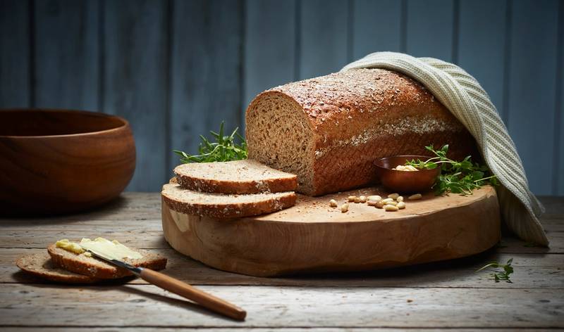 Et stort, grovt brød ligger på et skjærebrett med noen skiver skåret av, omgitt av en liten skål med smør og noen urter, på et mørkt trebord med en blå bakgrunn.