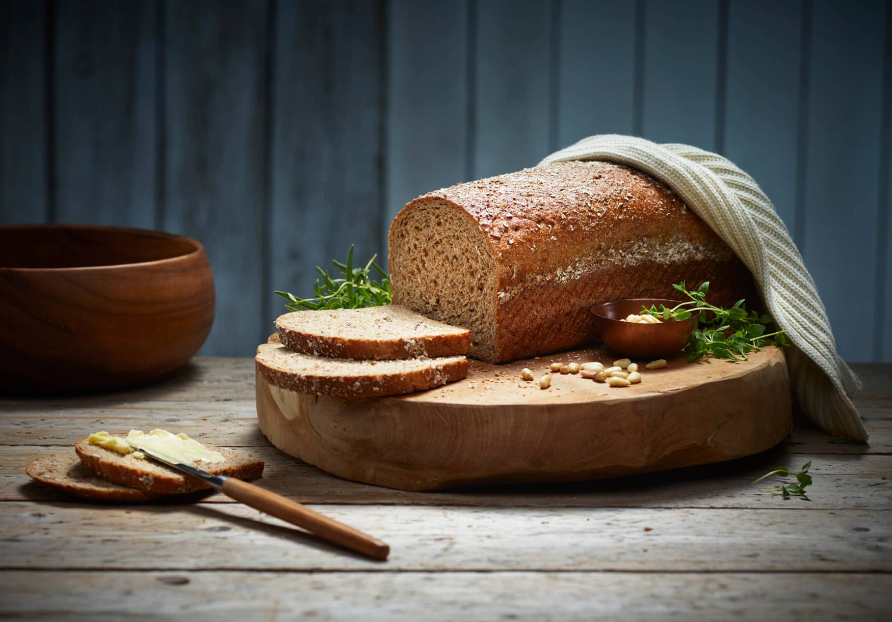 Et stort, grovt brød ligger på et skjærebrett med noen skiver skåret av, omgitt av en liten skål med smør og noen urter, på et mørkt trebord med en blå bakgrunn.