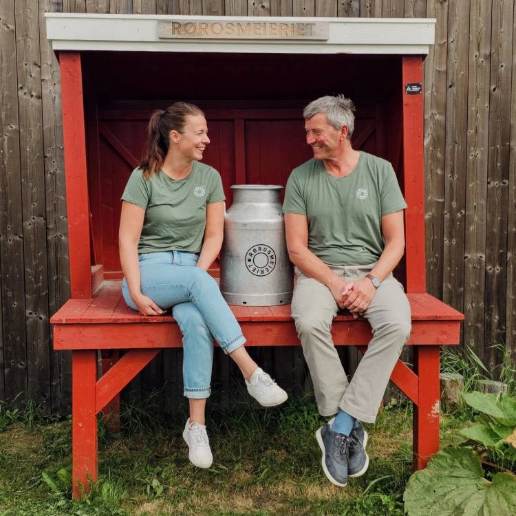 To personer sitter på en benk foran Rørosmeieriet, med en melkekanne mellom seg, i en utendørs setting med treverk og gress.