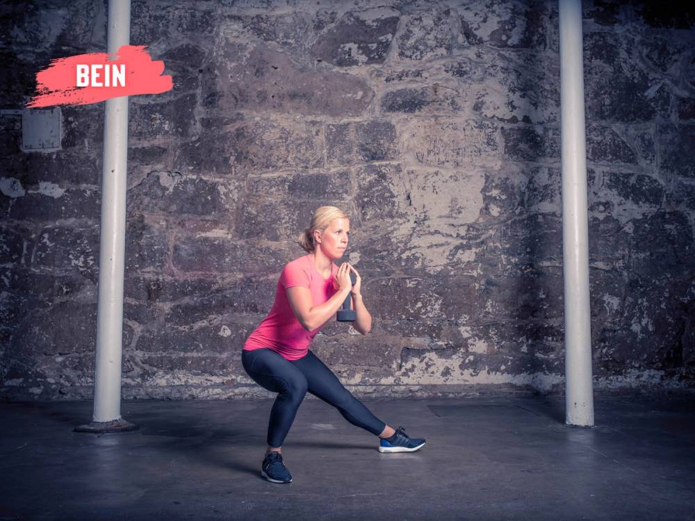 En person i rosa treningtøy gjør en knebøyøvelse foran en murvegg, med stangstativ på hver side.