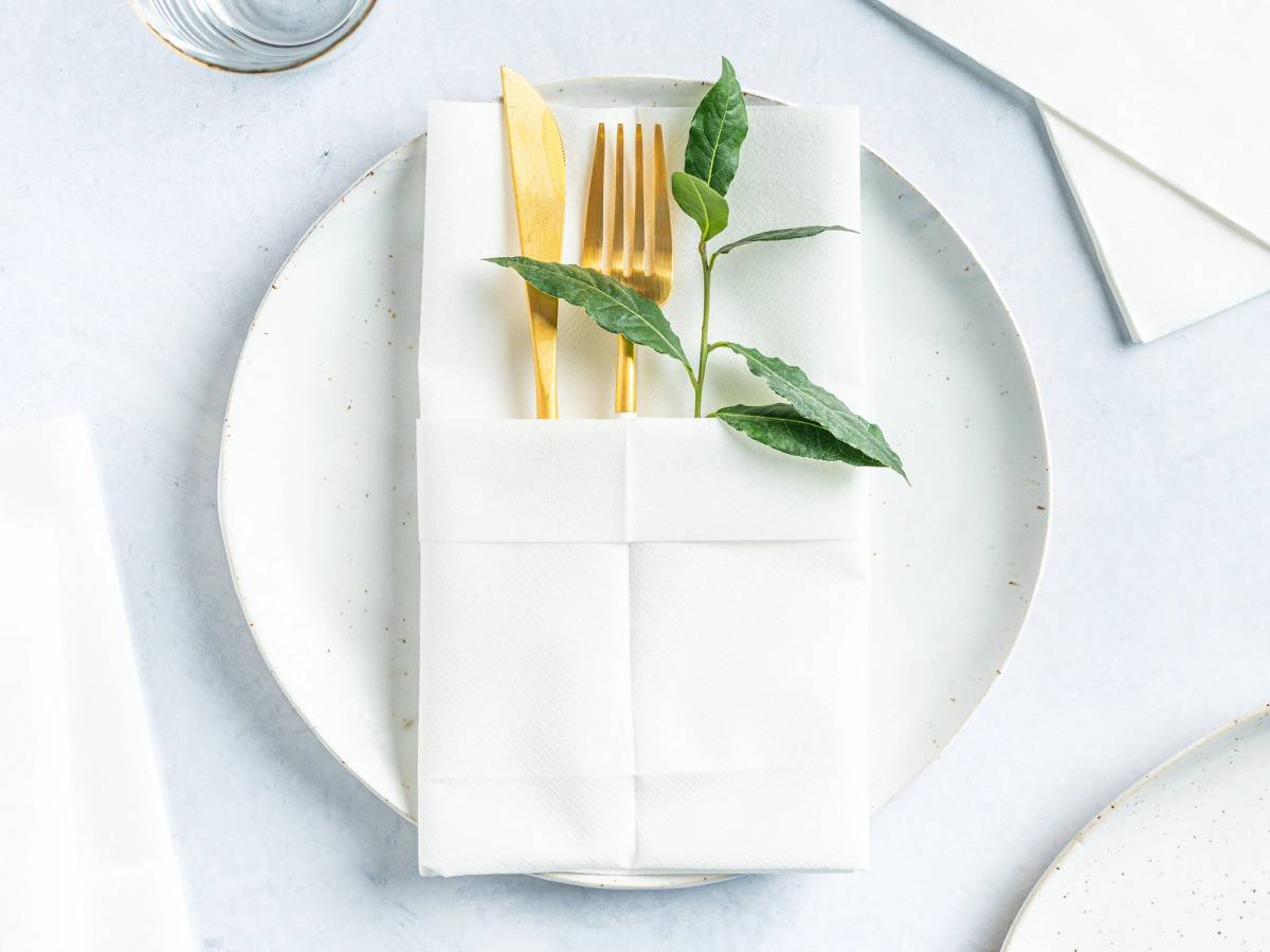En hvit serviett brettet rundt en gullfarget gaffel og kniv, pyntet med grønne blader, på en hvit tallerken.