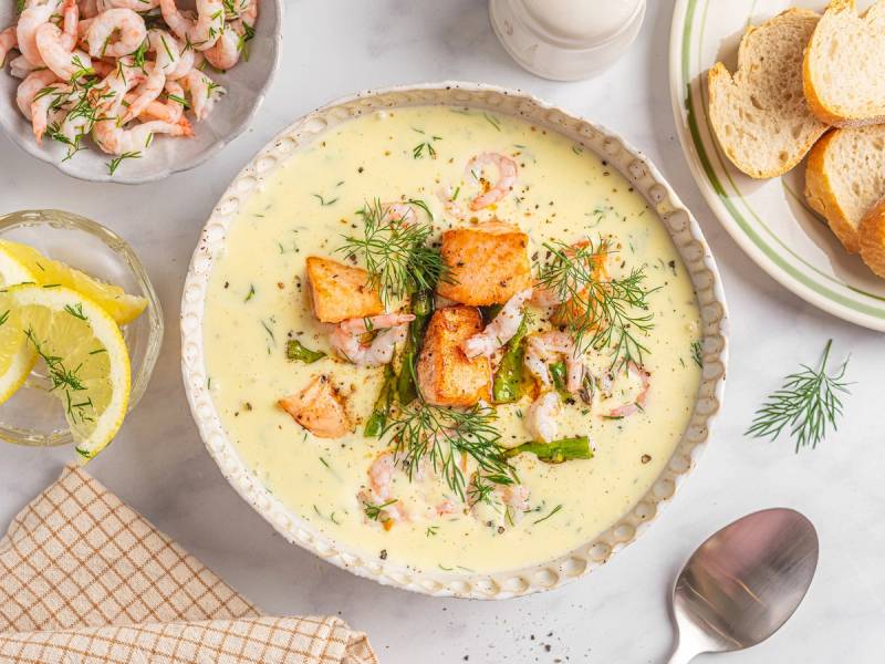 Kremet fiskesuppe med laks og reker servert i en skål, omgitt av brødskiver, sitron og dill, på et bord.
