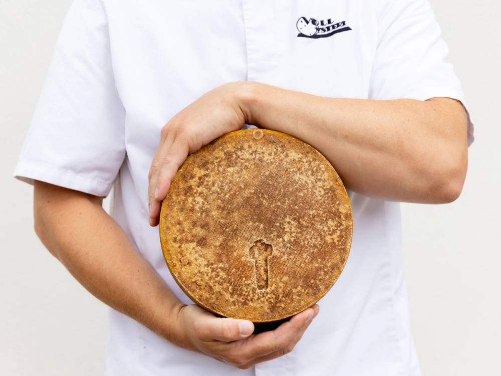 En person i hvit kokkeforkle holder en stor rund kake med en nøkkelformet skjult inntrykk i midten.