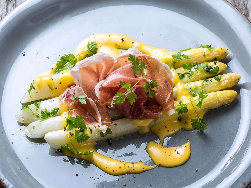 Kokte hvite asparges med hollandaise saus og skinke på en tallerken, pyntet med urter.
