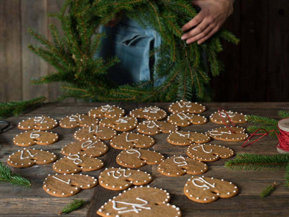 Pepperkakekalender med små pepperkaker formet som hjerter og andre figurer, dekorert med hvit glasur og tall, omgitt av granbar og en blå gavepose.