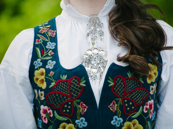 Nærbilde av en bunadsskjorte med broderte blomster og hjerter, og en sølvspenne i halsen.