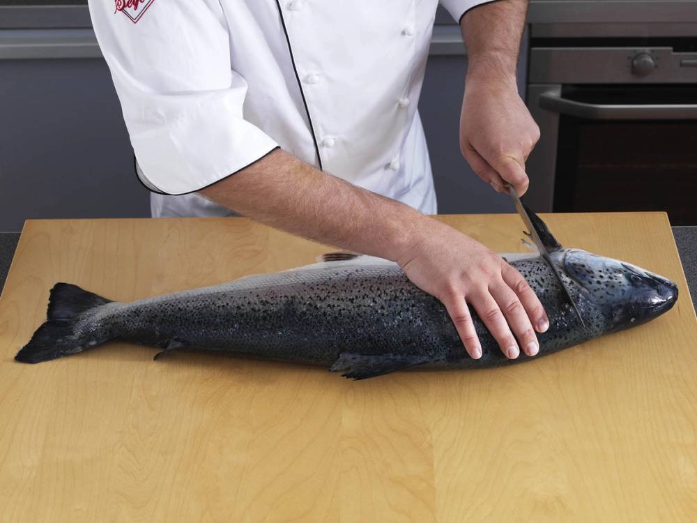 Person som fileterer en hel laks på et kjøkkenbord med en kniv.