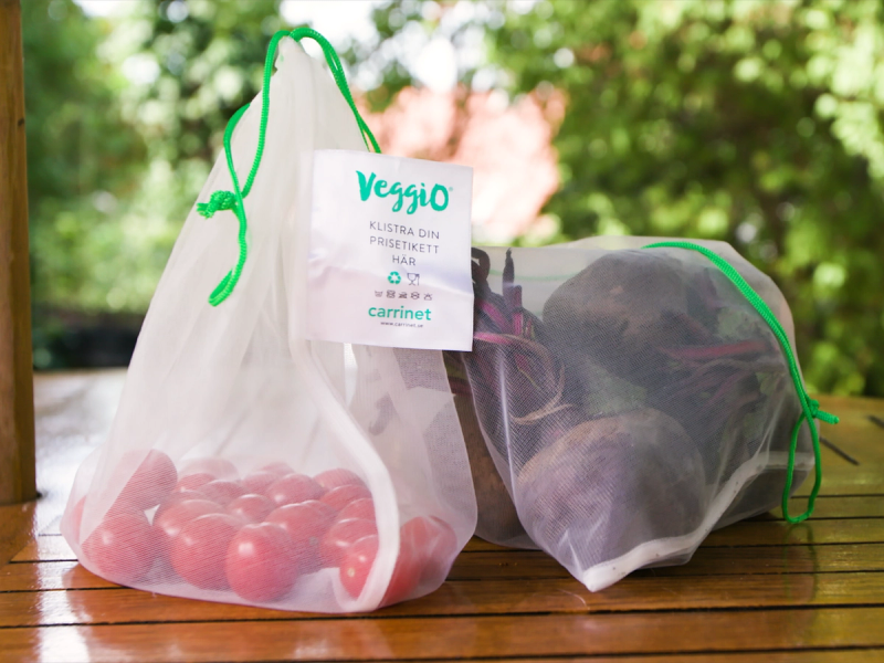 To gjenbruksposer, en med tomme tomater og en annen med noe mørkt innhold, ligger på et trebord utendørs med grønt løv i bakgrunnen.