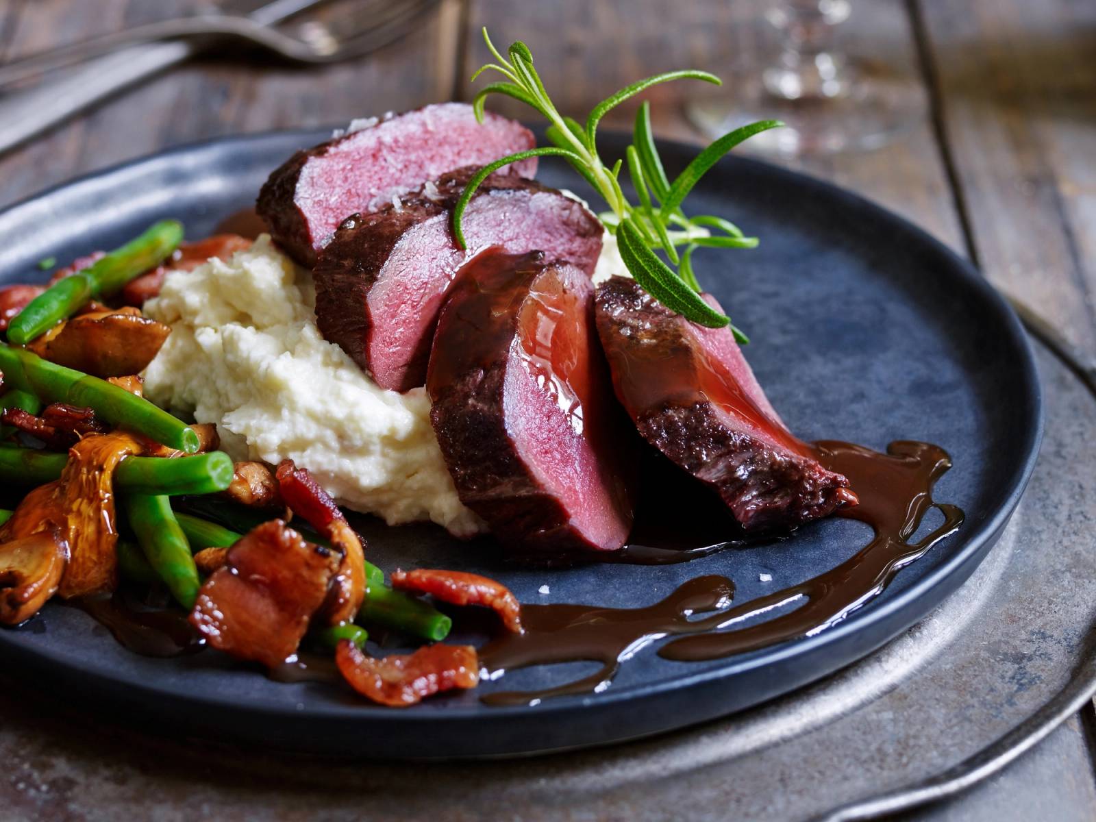 Renskav med saus, potetmos og grønnsaker på en svart tallerken.