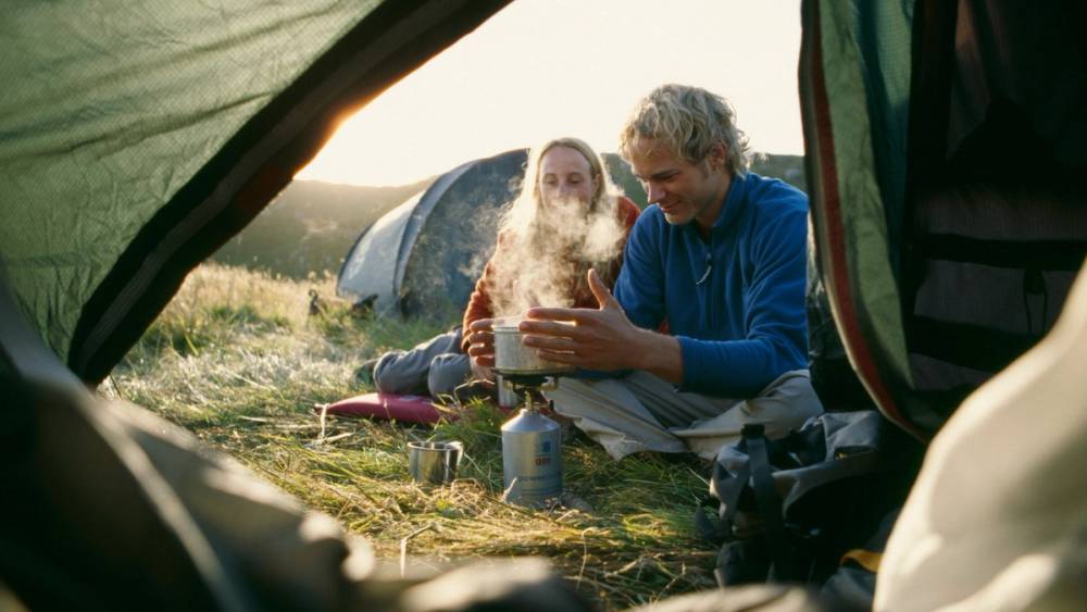 To personer sitter ved et bål i en campingplass, sett fra innsiden av en teltåpning, med natur i bakgrunnen.