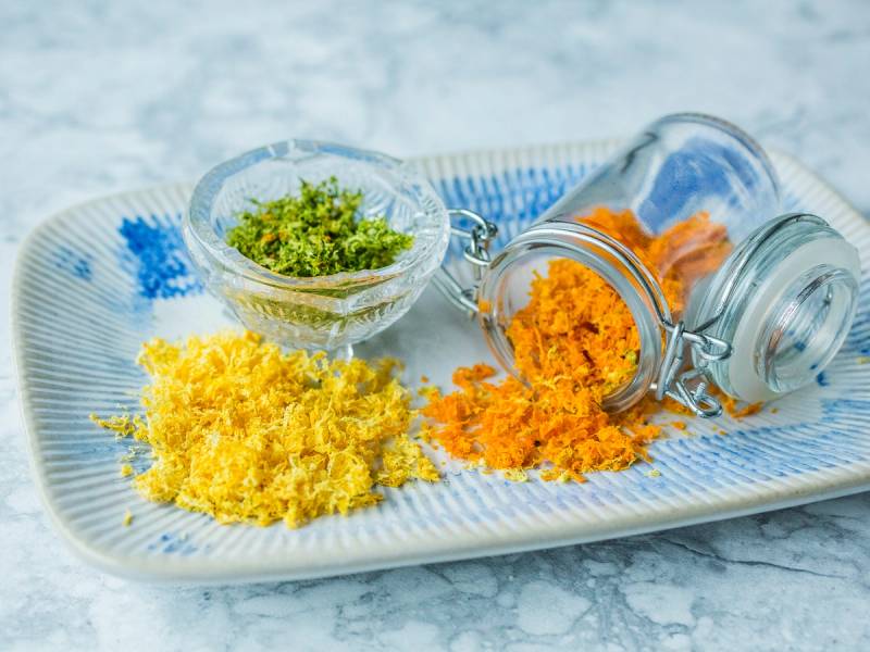 Tørkede sitrus-skall på en hvit tallerken med en liten skål med grønt krydder og et glass med tørkede sitrusbiter, sannsynligvis appelsin eller sitron, på et marmorbord.