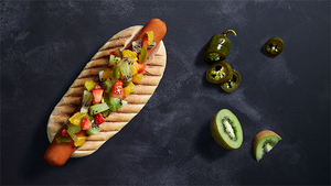 En pølse i brød med spesiell salsa og noen grønne chilipepper på en svart bakgrunn.