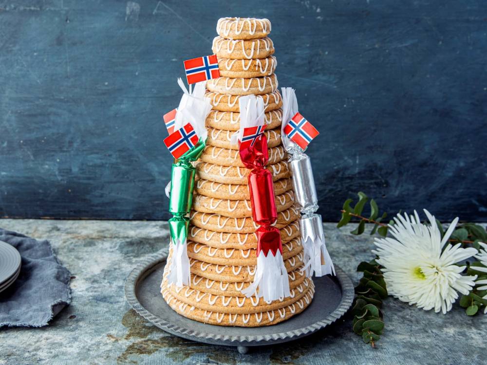Kransekake pyntet med norske flagg og små pakker, plassert på et fat med blomster i nærheten.