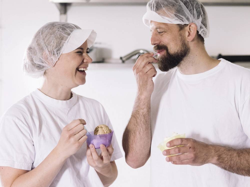 To personer i hvite klær og hårnett som smaker på iskrem i et kjøkkenmiljø, uten å vise ansiktene tydelig.