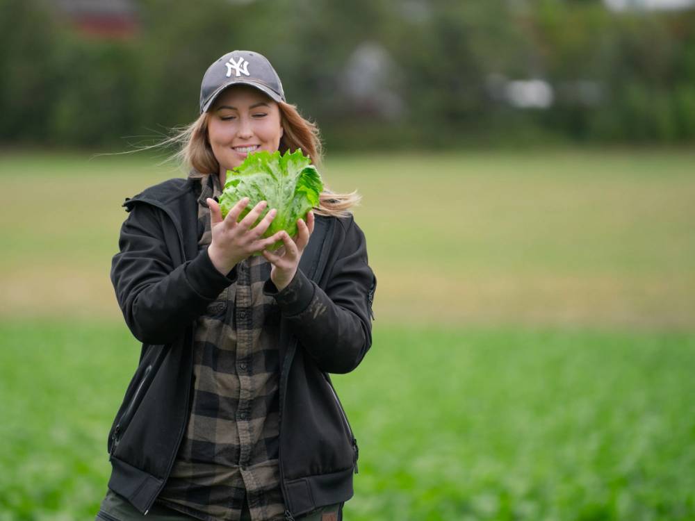 En person holder en stor salat med grønnsaker i en park, med trær og gress i bakgrunnen.