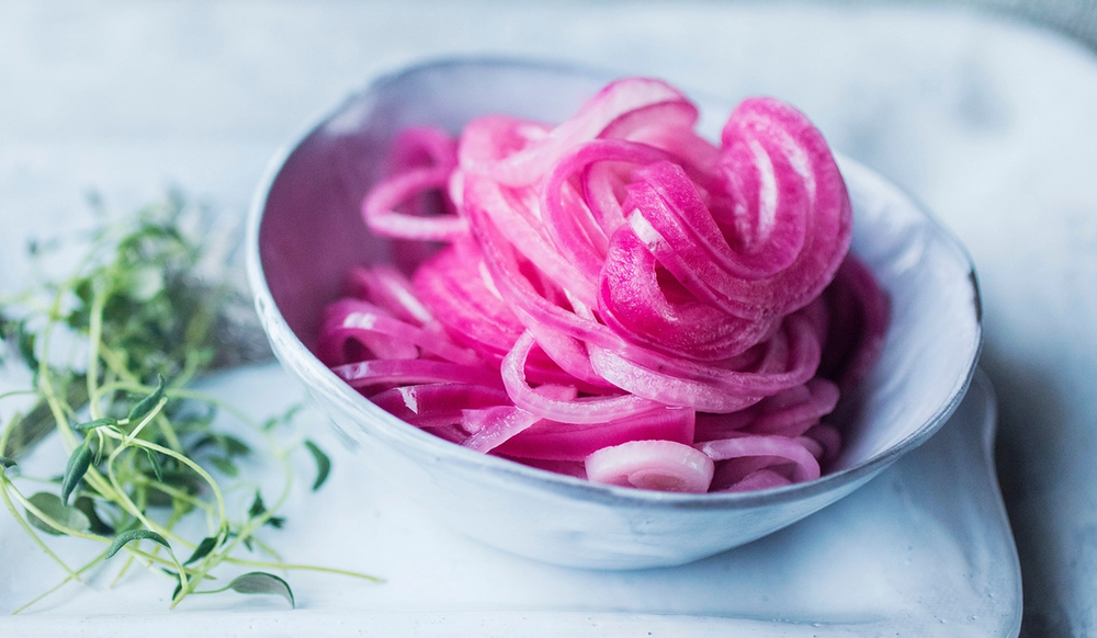 Skålen inneholder syltet rødløk med rosa farge, plassert på et hvitt fat med noen grønne urter ved siden av.