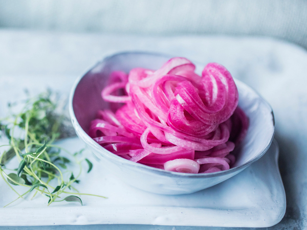 Skålen inneholder syltet rødløk med rosa farge, plassert på et hvitt fat med noen grønne urter ved siden av.