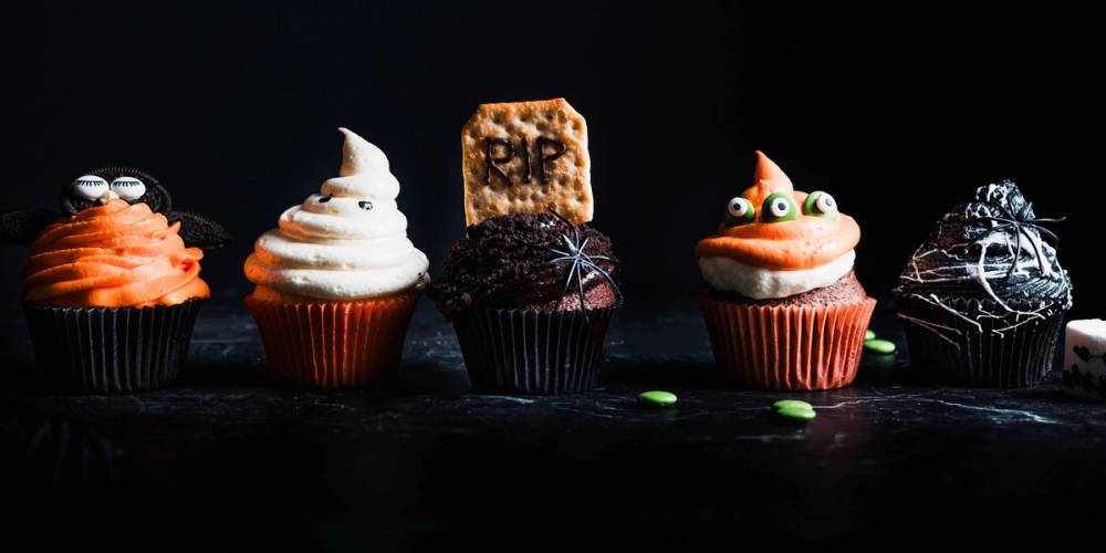 Fem halloween-inspirerte cupcakes med ulike dekorasjoner på en svart bakgrunn.
