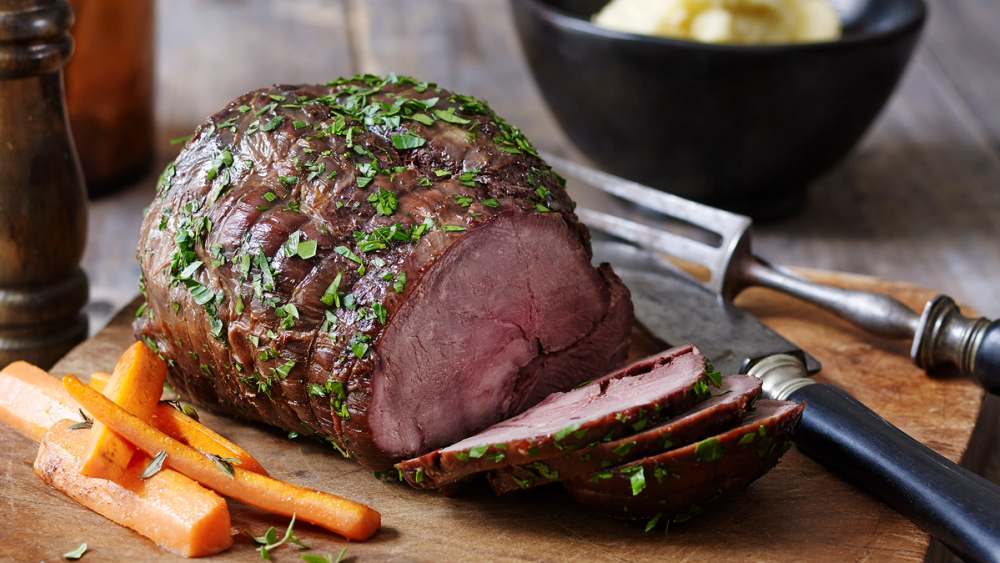 Svært saftig og mør stek av viltkjøtt med friske urter, skåret opp på et skjærebrett med grønnsaker og en kniv ved siden av.