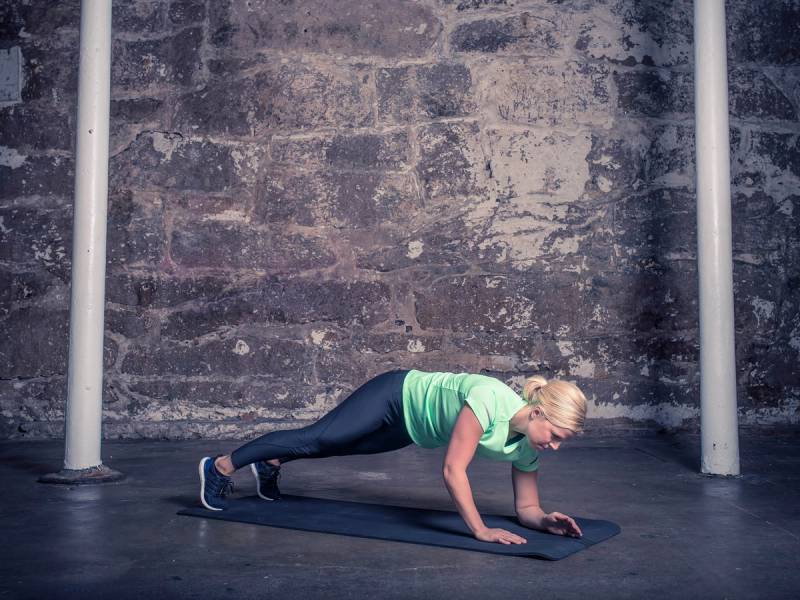 En kvinne gjør plankevandring på en treningsmatte i et treningsstudio med murvegg i bakgrunnen.