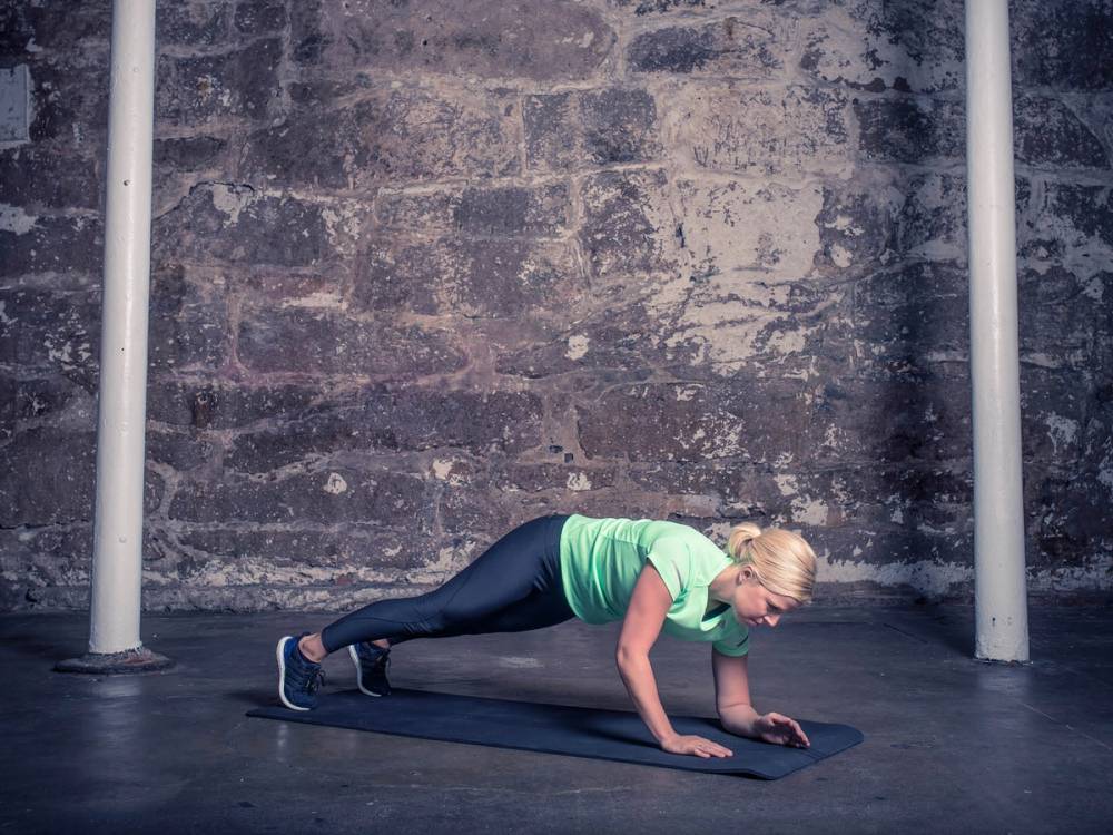 En kvinne gjør plankevandring på en treningsmatte i et treningsstudio med murvegg i bakgrunnen.