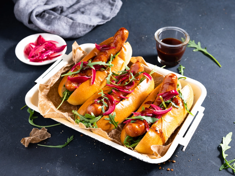 Tre pølser i brød med cheddar og chipotle, servert på en hvit tallerken med tilbehør og en liten skål med saus, på en mørk bakgrunn.