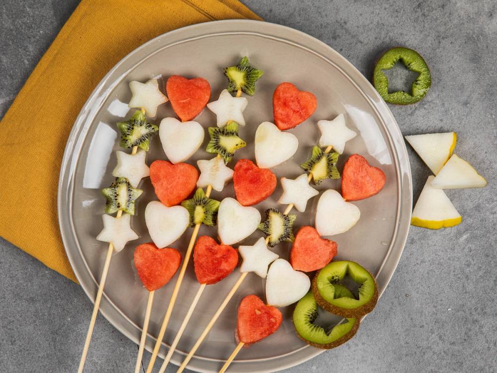 Skål med hjertedeiger og stjerner av frukt på tre spyd, omgitt av skiver av kiwi og eple