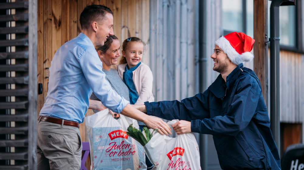 To personer utveksler gaver under juletreet, med en person som har på seg en nisselue, i en koselig innendørs setting.