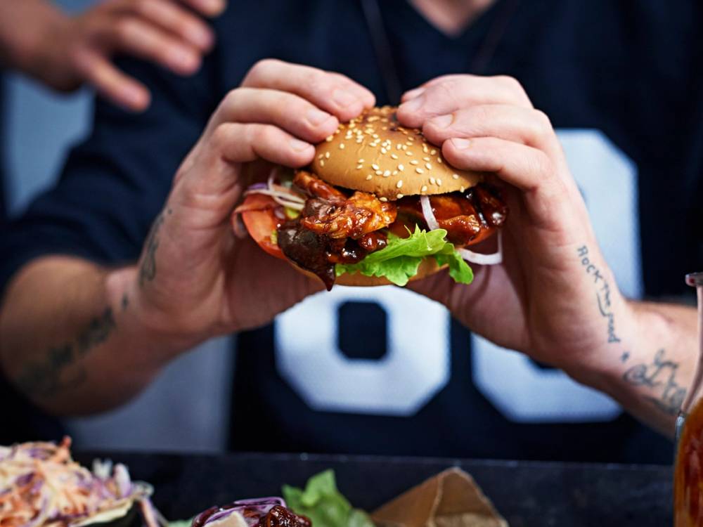 Person holder en burger med kjøtt, salat og saus i et brunt hamburgerbrød, klar til å spise.